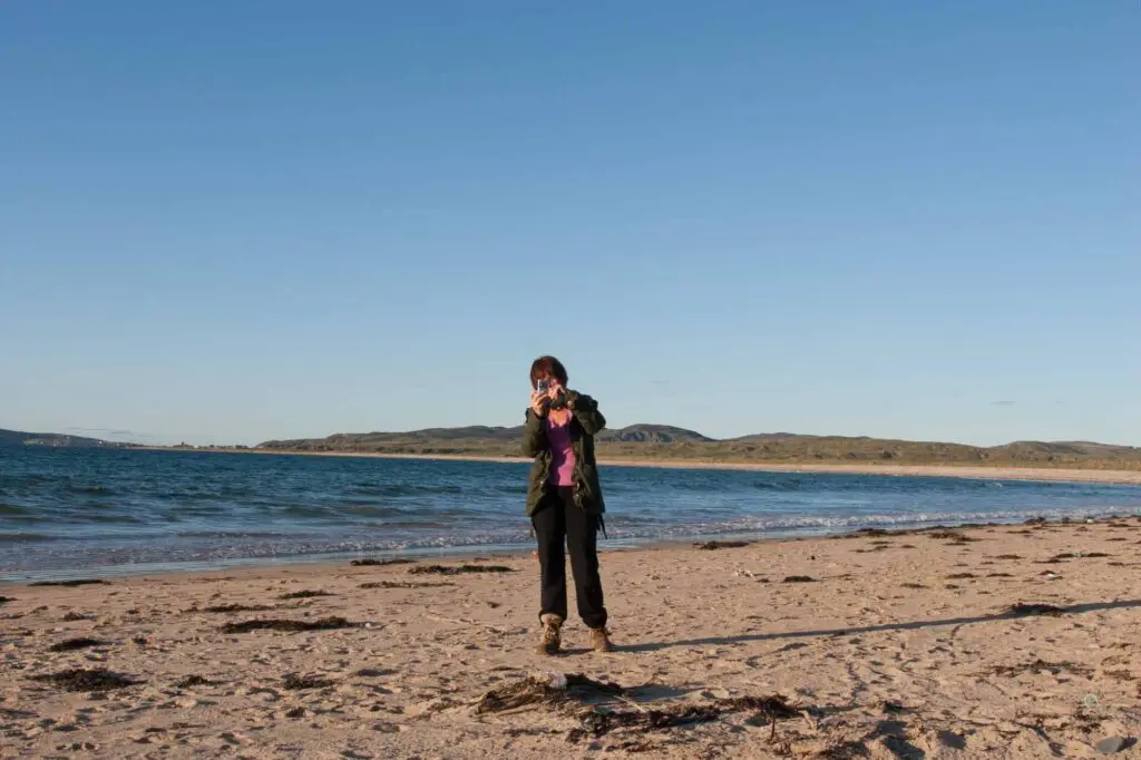 A woman taking a photograph on Ballyliffin Beach with sand dunes behind her and Carrickabraghy Castle in the far distance